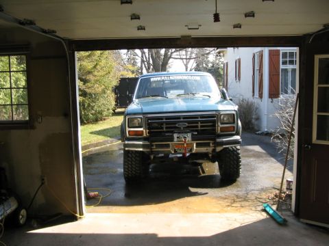 1986 Ford Bronco - A blue Ford Bronco is parked facing out of a garage, showing its front grille and large tires.