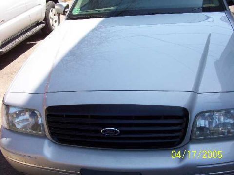 1999 Ford Crown Victoria - The front of a silver Ford Crown Victoria is visible, with a black grille and headlights, alongside another vehicle.