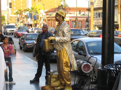 1968 Ford Mustang - A street performer in a gold costume stands on a sidewalk with onlookers and traffic in the background.