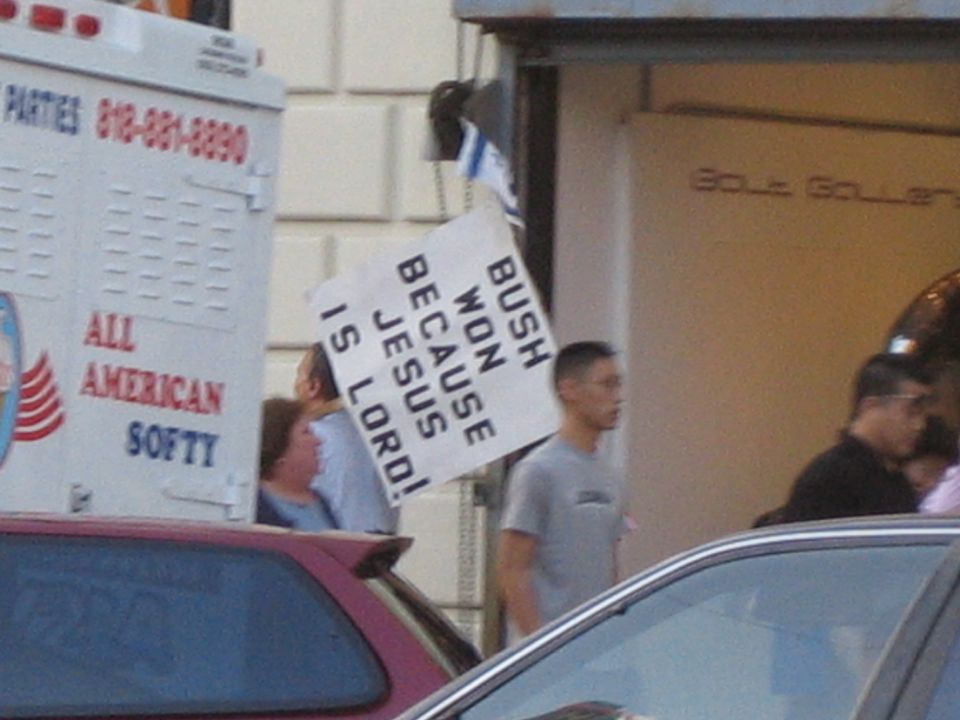 People stand near an ice cream truck and cars, one holding a sign that reads 'BUSH WON BECAUSE JESUS IS LORD!'
