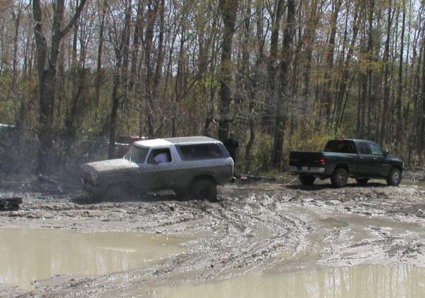 Mud-caked Ford Bronco buried to the rockers in a muddy pit, dark pickup parked at the treeline