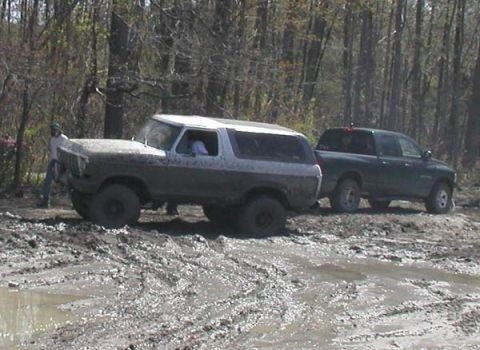 1988 Ford Bronco - Two trucks navigate a muddy terrain with trees in the background.