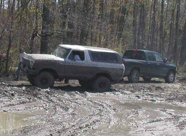 Mud-covered 1980s Ford Bronco with a white top, stuck in mud next to a dark green pickup truck
