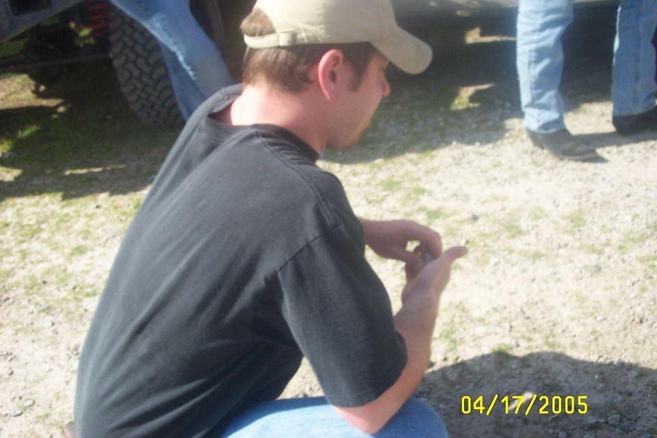 guy in black shirt and tan hat crouches near a vehicle tire, other people stand nearby. Date stamp reads 04/17/2005