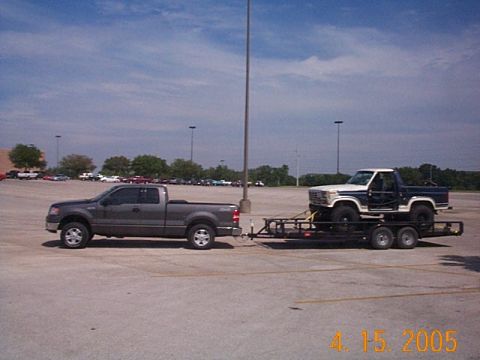 1986 Ford Bronco - Pickup truck tows a trailer carrying a classic Ford Bronco. The Bronco appears to be from the early 1980s.