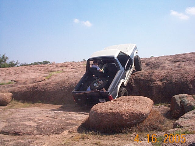 Dark gray and white Ford Bronco climbing large rocks, driver visible in cab