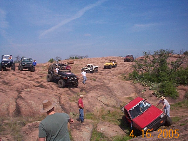 Red off-road vehicle tilted on rocks with people around, other SUVs on hill