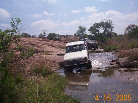 1986 Ford Bronco - Multiple off-road vehicles navigate a rocky stream crossing in a natural landscape.