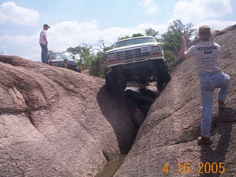 1986 Ford Bronco - Two trucks are navigating a challenging off-road course with steep rock inclines and water below.