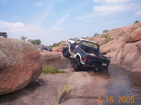 1986 Ford Bronco - A Ford Bronco navigates a rocky stream bed, with other vehicles visible in the background.