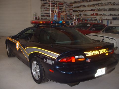 1999 Ford Crown Victoria - This dark blue police car, marked "HIGHWAY PATROL", is parked in a garage with other vehicles and shelves of trophies.