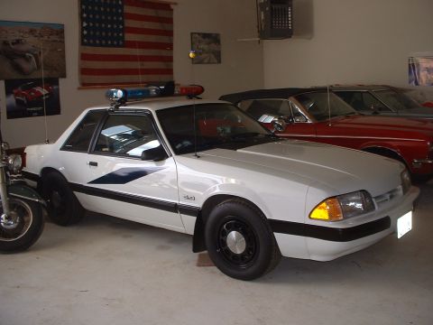 1999 Ford Crown Victoria - A white police car is parked in a garage next to other vehicles and an American flag.