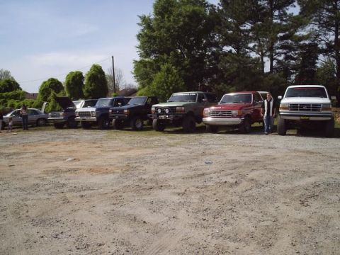 1989 Ford Bronco - A lineup of Ford trucks and SUVs are parked in a dirt lot under a clear sky.