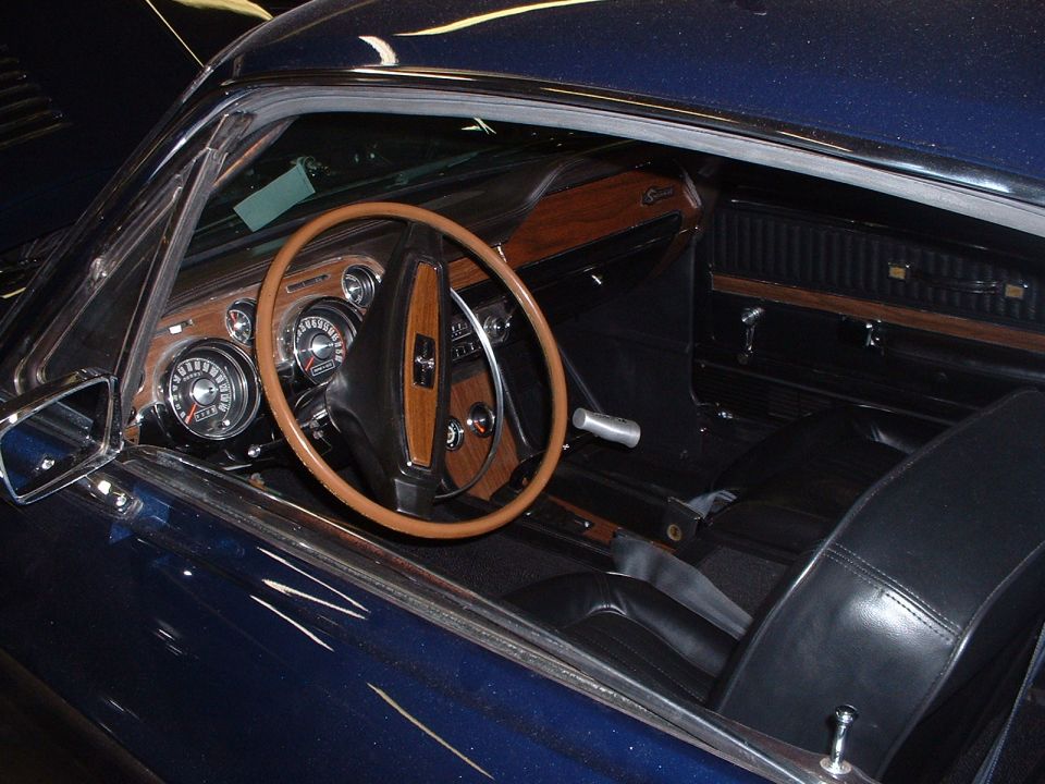 Dark blue car interior with wood trim, wood-rimmed steering wheel, and black leather seats. Likely a 1968 Mustang