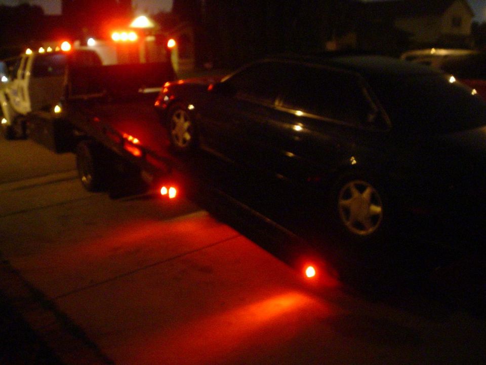 Dark sedan being towed at night by a tow truck with red lights