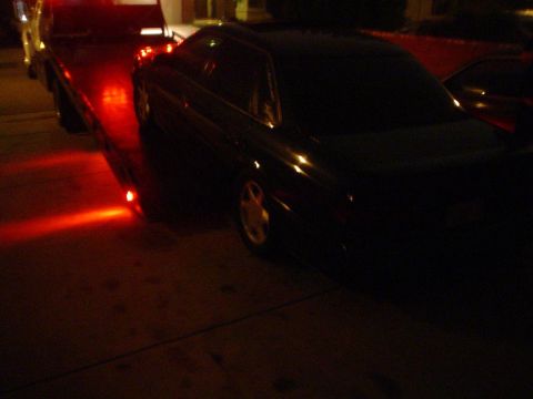 1993 Ford Taurus - A dark 1993 Ford Taurus is loaded onto a tow truck at night, illuminated by red lights.