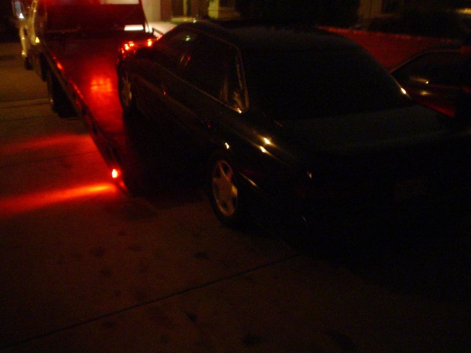 Dark 1993 Ford Taurus on a flatbed tow truck at night, viewed from the rear quarter