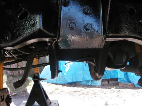 1994 Ford Bronco - Underbody view of a truck raised on a jack stand, looking up at freshly painted black frame rails, a boxed crossmember and the transmission…