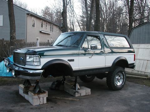 1994 Ford Bronco - A two-tone Ford Bronco is elevated on jack stands in a driveway, showing its front end.