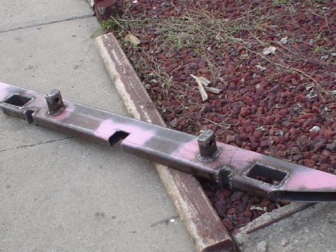 1981 Ford Bronco - A fabricated steel bumper laid diagonally across a driveway edge and lava-rock bed, its face mottled with pink primer and ground-bare metal…