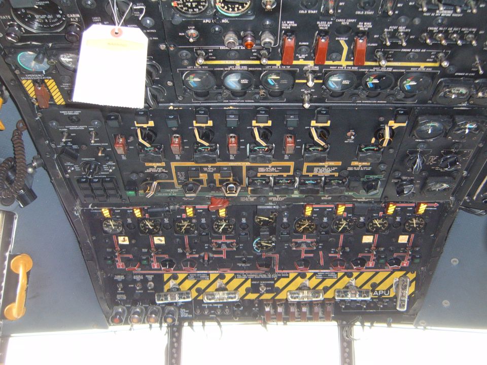 Aircraft cockpit instrument panel with many gauges, switches, and labels. A white tag hangs from the top