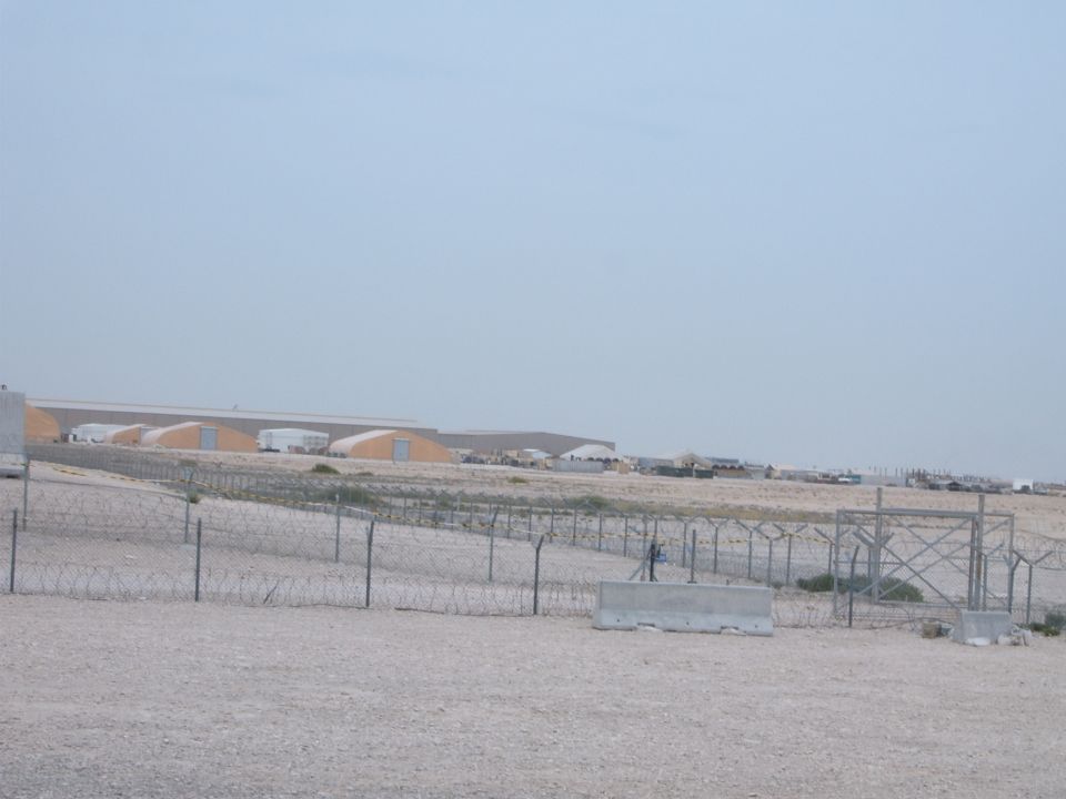 Desert landscape with barbed wire fence, concrete barriers, and industrial buildings under a hazy sky