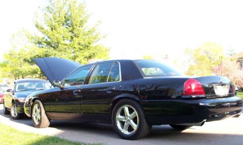 1998 Ford Crown Victoria - Two cars parked outside, one with its hood up, on a sunny day.