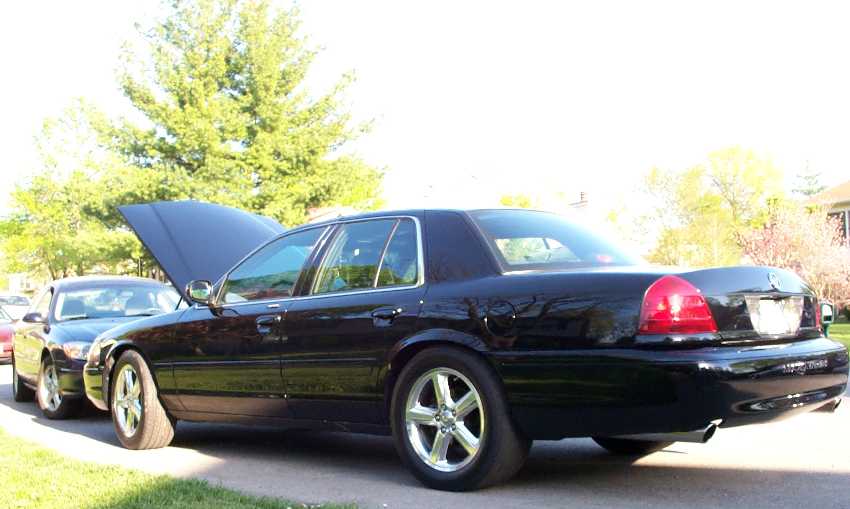 Black full-size sedan, rear 3/4 view, hood up, polished wheels, in driveway