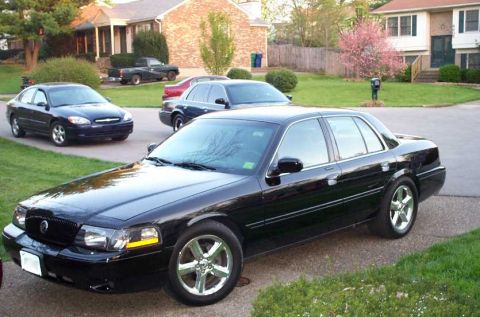 1998 Ford Crown Victoria - A black sedan is parked in a suburban driveway with other vehicles and houses visible.