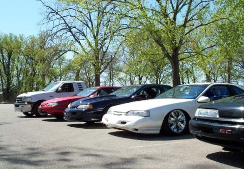 1998 Ford Crown Victoria - A row of vehicles parked at an angle in a paved lot under leafy trees, including a white pickup truck, a red car, a dark blue coupe, a whit…