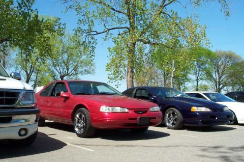 1998 Ford Crown Victoria - A red Ford Thunderbird coupe parked in a gravel lot alongside a dark blue Ford Mustang coupe, with a white SUV in the foreground left, all…