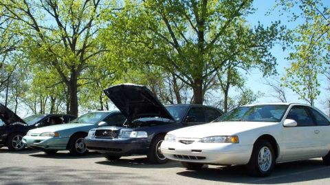 1998 Ford Crown Victoria - A row of cars parked under leafy trees, featuring a dark blue Ford Crown Victoria sedan with its hood open in the center, flanked by a whit…