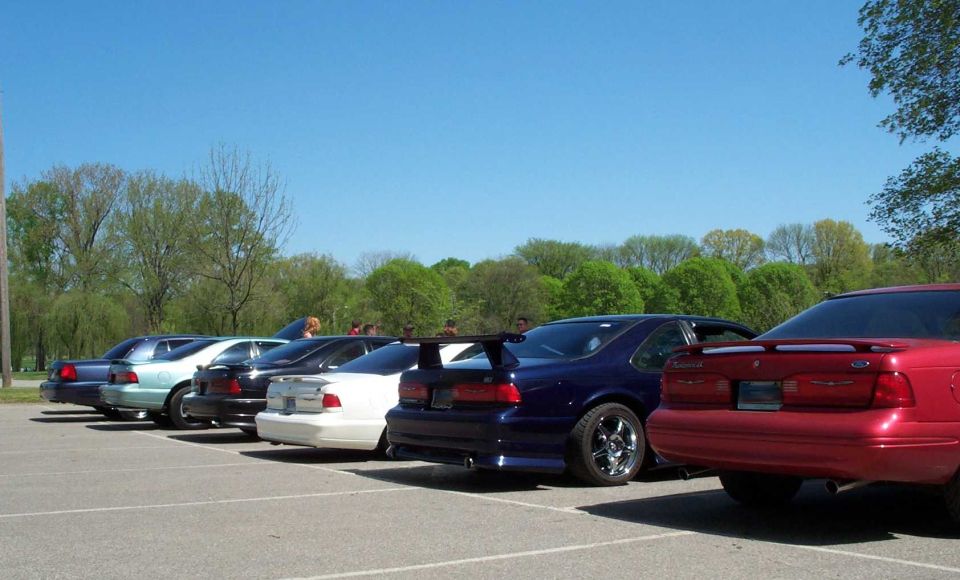 Row of six 1990s Ford Thunderbird coupes parked rear-on in a sunny lot, one with a tall aftermarket wing