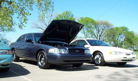 1998 Ford Crown Victoria - A dark blue/gray Ford Crown Victoria sedan sits in a paved parking lot with its hood raised, exposing the engine bay, chrome grille with Fo…