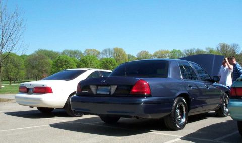 1998 Ford Crown Victoria - A dark blue/gray Ford Crown Victoria sedan viewed from the rear three-quarter angle with its trunk open, parked in a paved lot next to a wh…