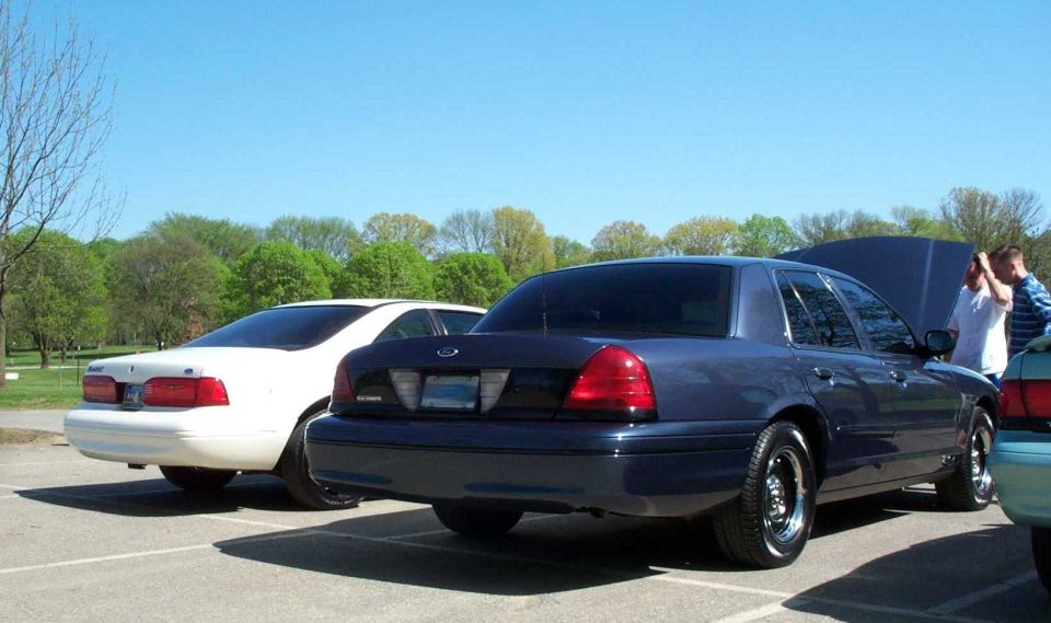 Dark blue Ford Crown Victoria sedan, rear 3/4 view with open trunk, beside a white sedan in a parking lot.