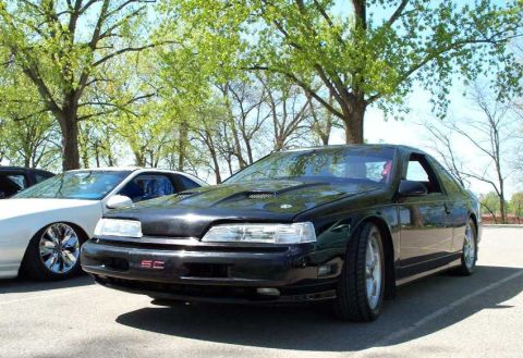 1998 Ford Crown Victoria - A black two-door coupe photographed from a front three-quarter angle, with a hood scoop, aftermarket multi-spoke wheels, and a red 'SC' emb…