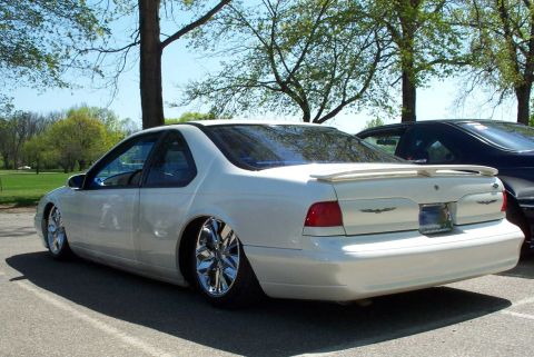1998 Ford Crown Victoria - White Ford Thunderbird coupe, lowered on chrome wheels with rear spoiler, parked in a tree-lined lot, rear three-quarter view.