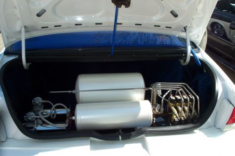 1998 Ford Crown Victoria - Open trunk of a white sedan showing a custom air suspension setup with two silver air tanks, dual black air compressors on the left, and a…