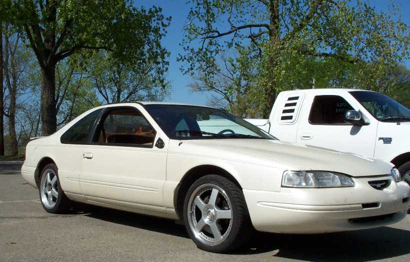 Cream two-door coupe, 3q front view, with white pickup behind under trees