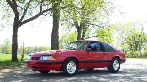 1998 Ford Crown Victoria - A red Fox-body Ford Mustang hatchback coupe with a '5.0' fender badge, viewed from the front-three-quarter angle, parked on pavement in a g…