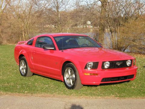 2005 Ford Mustang - A red Ford Mustang coupe is parked on a grassy area near some trees and a body of water.