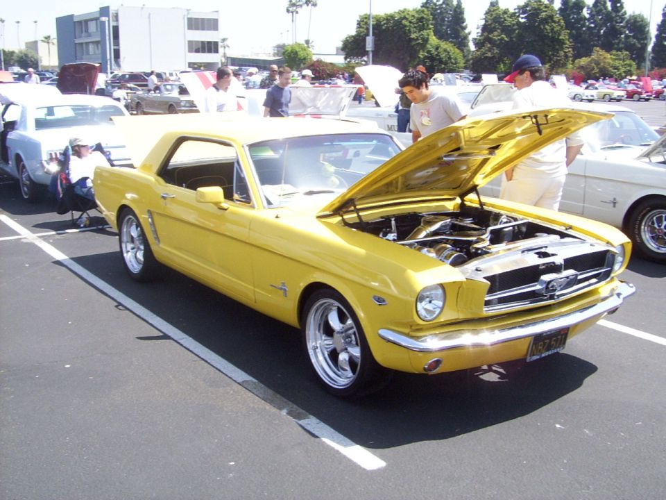 Yellow 1965 Ford Mustang coupe with hood open, chrome wheels, parked in a lot