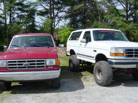 1988 Ford Bronco - Two Ford Broncos are parked side-by-side, one red and one white with large tires.