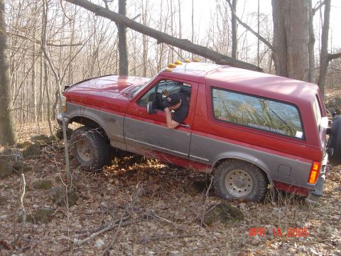 1995 Ford Bronco - A red and grey Ford Bronco is seen off-roading through a wooded area with rocky ground.
