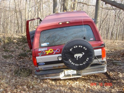 1995 Ford Bronco - A red Ford Bronco is parked off-road in a wooded area, with its spare tire cover prominently displayed.