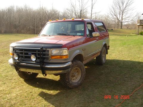 1995 Ford Bronco - This red and silver 1995 Ford Bronco is parked on a grassy lawn with trees in the background.