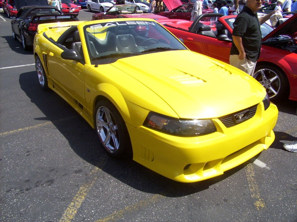 Yellow convertible with a Saleen sticker on the windshield, parked at a car show. Other cars are visible in the background
