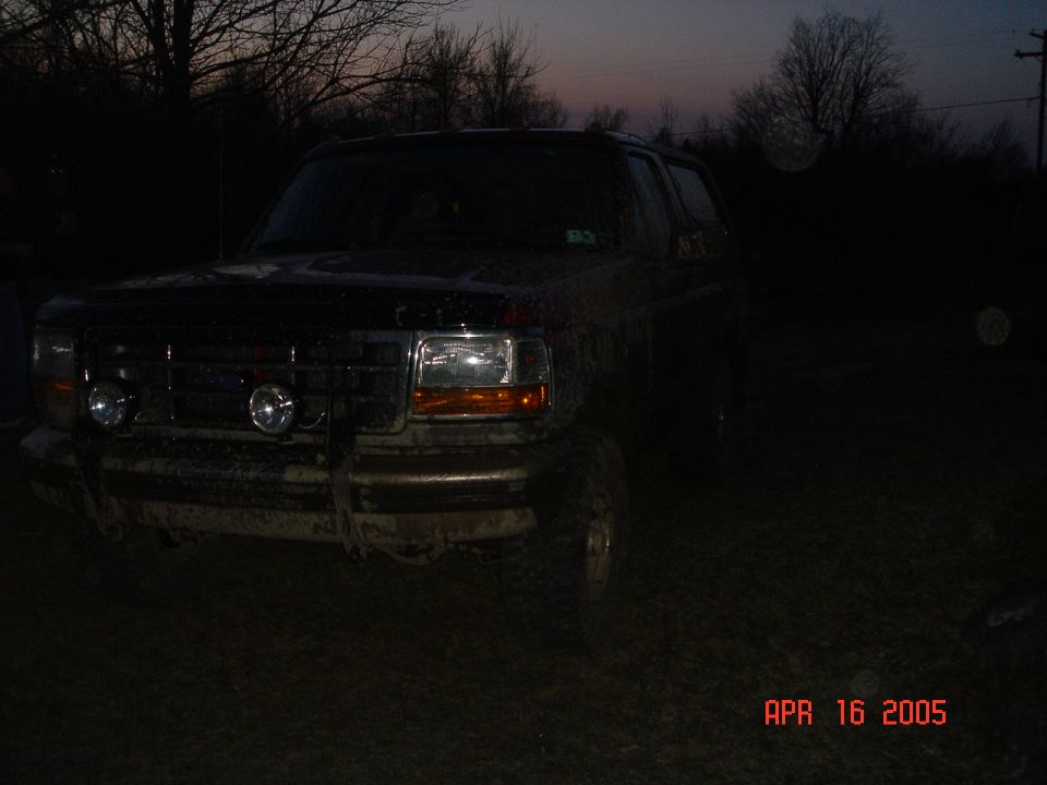 Mud-covered lifted Ford Bronco with brush guard and light bar at dusk in a field