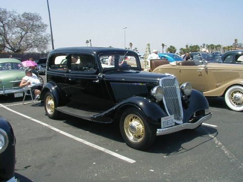 1993 Ford Probe - Black 1934 Ford Model 40 sedan with gold wheels at a car show, other vehicles in background.